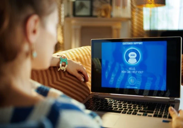 A woman sitting on a couch with a virtual customer support agent on a laptop.