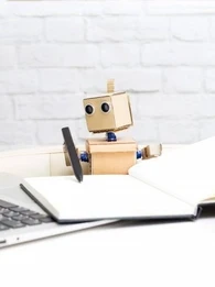 Toy robot pretending to write in a notebook while looking at a laptop