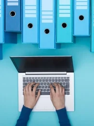 A person working on a laptop with large files and folders hanging above
